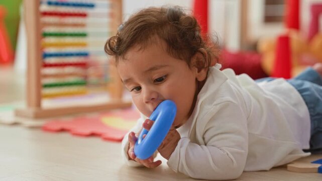 Girl mouthing blue ring on mat in playroom with abacus and cones nearby; curiosity discovery early learning.