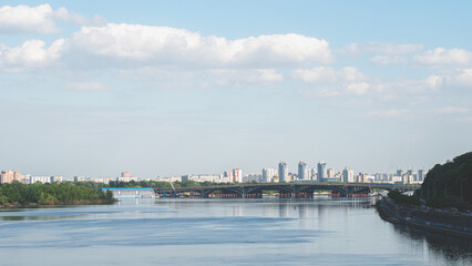 Naklejka premium Kyiv city skyline view across the Dnieper River featuring the Metro Bridge, water reflections, and modern residential buildings under a cloudy sky in Ukraine.