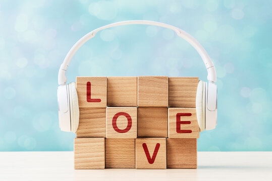 Word LOVE on wooden cubes with white headphones on a blue bokeh background. Creative music concept banner representing love for audio content, podcast and romance.