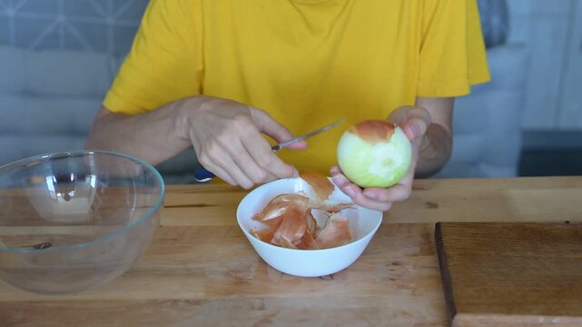 Woman peeling yellow onion with knife above bowl at wooden table in bright home kitchen video Full HD