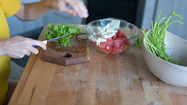 Woman chopping fresh herbs beside tomato and tofu salad bowl on wooden table in home kitchen video Full HD