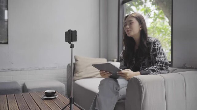 A young woman sits on a sofa in a sunlit living room, speaking and gesturing toward a smartphone mounted on a tripod while holding a tablet