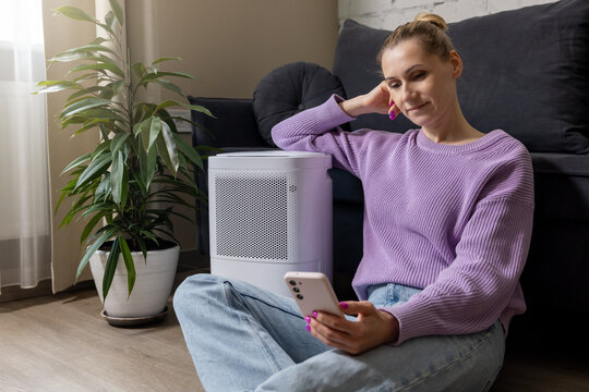 woman sitting on the floor by air purifier in living room and using mobile phone. air quality and microclimate control at home