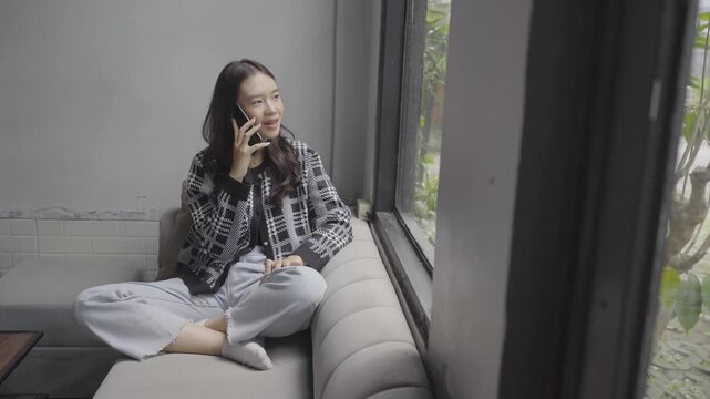 Throughout the clip, a young woman sits cross-legged on a sofa beside a large window, first looking at her phone, then talking, and finally smiling slightly