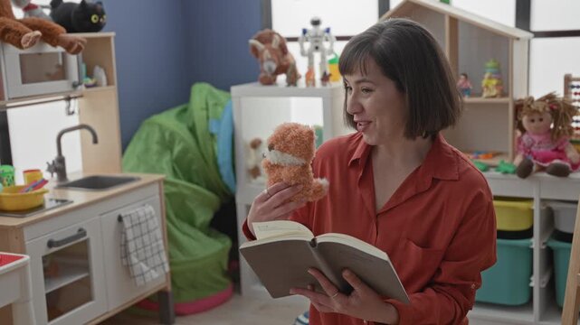 Woman reading book with puppet, holding orange stuffed toy and smiling while gesturing to toys in a preschool building; joyful engagement.