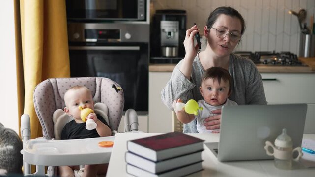 Mother multitasking with laptop phone babies in kitchen working from home. Woman works on computer while baby sits in highchair. Mom balances work with childcare. Parent manages remote work.