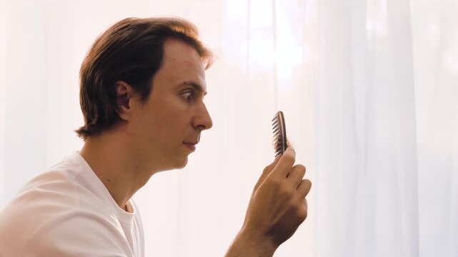 A shocked man finds fallen hair on his comb while grooming. This portrait highlights male hair loss, stress-related alopecia, and balding concerns in the morning light. High quality 4k footage
