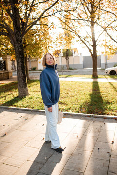 Stylish smiling senior woman 55-60 year old wearing blue sweatshirt and denim pants holding tote bag over sunny autumn background in park. 60s.