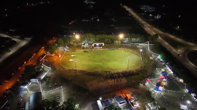 Vista a&eacute;rea de fiesta gaucha nocturna con p&uacute;blico y pista ecuestre, carrera de barriles en el campo argentino