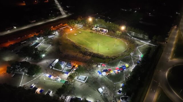 Vista a&eacute;rea de fiesta gaucha nocturna con p&uacute;blico y pista ecuestre, carrera de barriles en el campo argentino