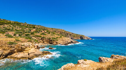 Fototapeta premium Stunning Mediterranean coastline featuring turquoise blue sea, rugged limestone cliffs, crashing white waves and traditional stone houses on a sunlit hillside