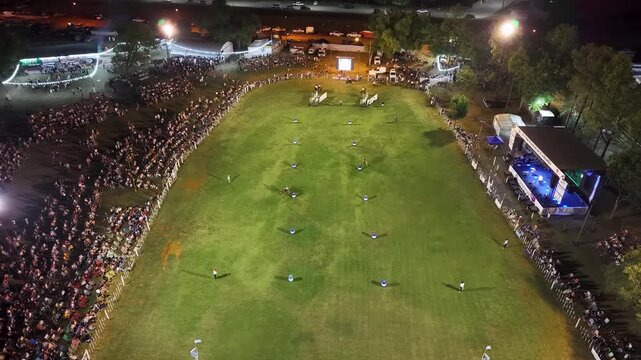 Vista a&eacute;rea de fiesta gaucha nocturna con p&uacute;blico y pista ecuestre, carrera de barriles en el campo argentino