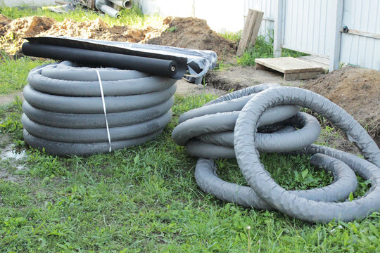 A coiled corrugated drainage pipe bundle lies on grass near a construction or landscaping area, with soil piles and a pale fence in the background.