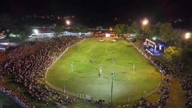 Vista a&eacute;rea de fiesta gaucha nocturna con p&uacute;blico y pista ecuestre, carrera de barriles en el campo argentino