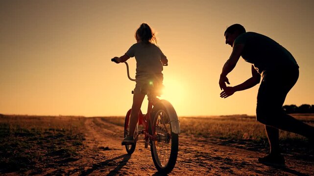 Parent teaching child to ride bicycle at sunset on dirt road in countryside. Adult runs beside young girl learning to cycle. Parent helps kid with bike during golden hour outdoor lesson.