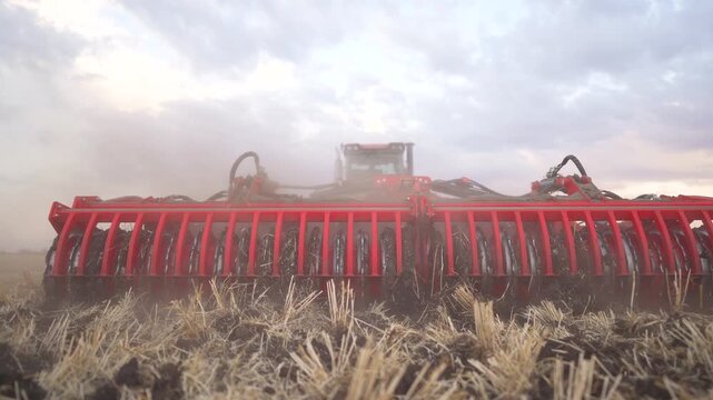 Red disc harrow tractor cultivating field with dry stubble under dramatic cloudy sky. Modern farm machinery working on agricultural field. Heavy tractor tilling and preparing soil for planting season.