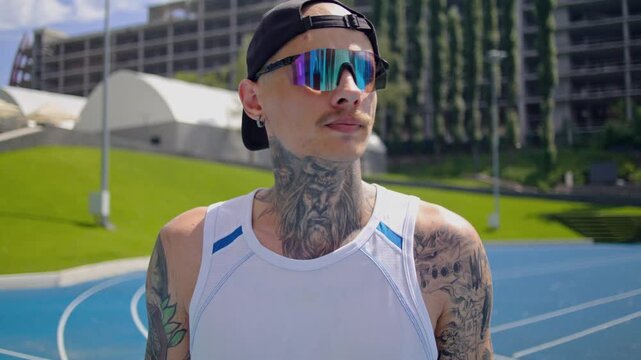 Tattooed male jogger with polarized sunglasses stands at stadium