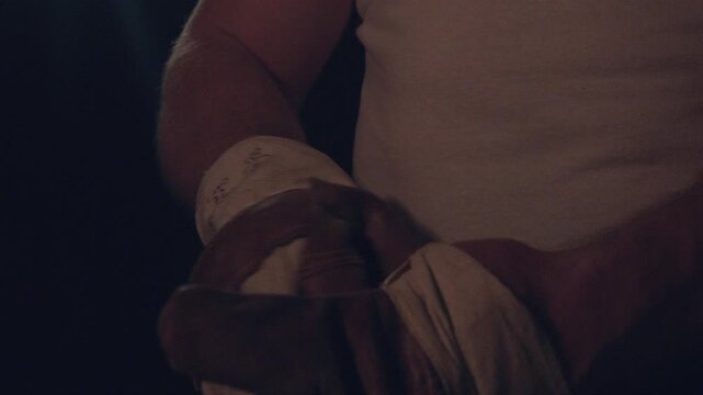 Muscular athlete wrapping hands with tape in dim gym, white tank top, focused ritual, wrist support, trainer guidance implied,