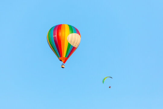 Masovian Voivodeship, Poland - 07 August 2018: View of a colorful rainbow-striped hot air balloon and a paraglider flying in a clear blue sky.