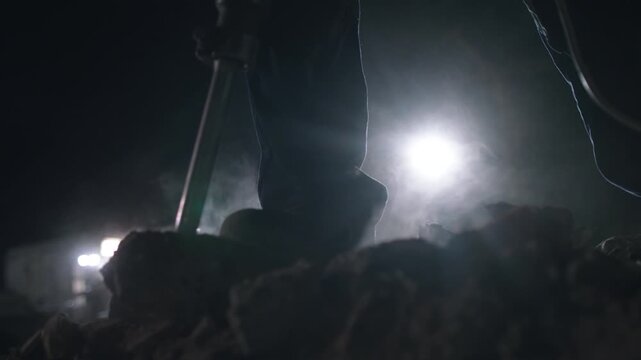 Lowangle illuminated ground drilling at night, tripod rig and rotating auger penetrate soil as bright lamp silhouettes operator