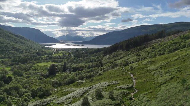 Pull-back drone footage reveals a winding path on a green hillside leading to the Pap of Glencoe. A village and loch are below under a cloudy sky in the Highland, Scotland, UK, on a spring afternoon.