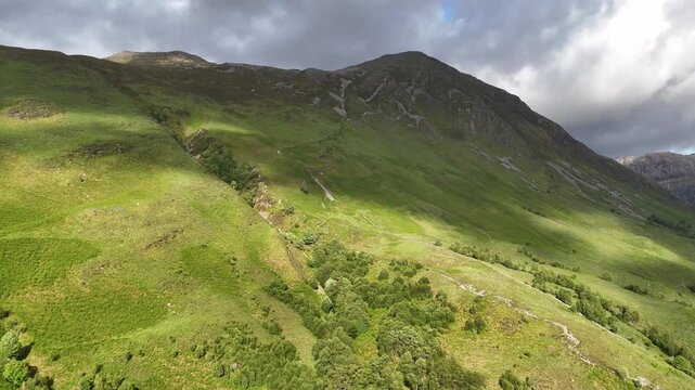 Push-in, boom up drone footage reveals a winding path on green slopes leading to the Pap of Glenco in the Highland, Scotland, UK, during a spring afternoon.
