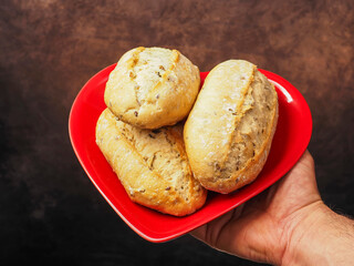 Naklejka premium Hand holds red heart shaped plate with three small sourdough rolls, grey painted background. High quality and value bread. Healthy bakery product. Love food concept. Ideal for food theme.