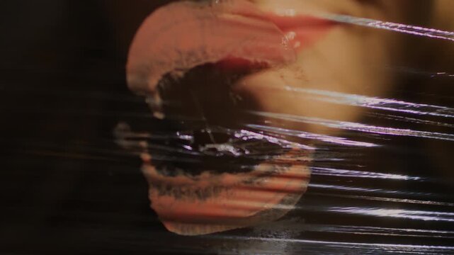 Close-up of woman's teeth biting on a screen of plastic wrap and leaving behind lipstick marks