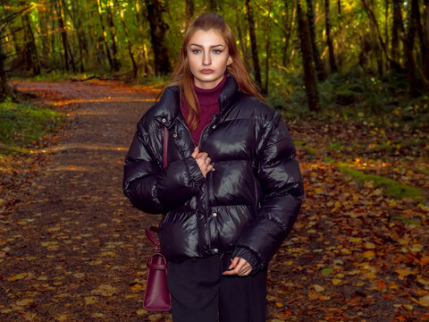 Woman in glossy black puffer jacket stands in autumn forest park. Sunlight filters through tall trees behind her. She gazes directly at the camera, calm and confident. Explore nature theme.