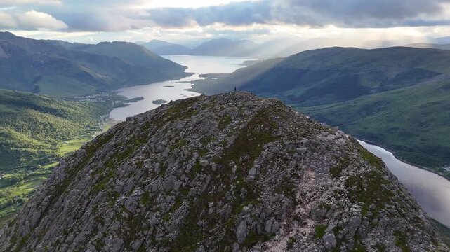 This aerial drone footage is a tracking shot that pushes forward towards a lone person standing on the rugged rocky peak of  Pap of Glencoe in Scotland UK.