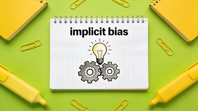 White spiral notebook with text implicit bias and a drawing of a lightbulb and gears on a bright green background with yellow office stationery supplies