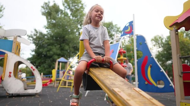 Happy girl rides seesaw at outdoor playground. Child laughs and smiles on teeter-totter in park. Girl enjoys playground fun with colorful equipment. Joyful child plays on seesaw outdoors.