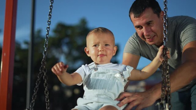 Happy father pushes baby on wooden swing. Father and child enjoy play at outdoor playground. Baby smiles during fast swing play. Family spends time in sunny park. Loving father holds baby safe.