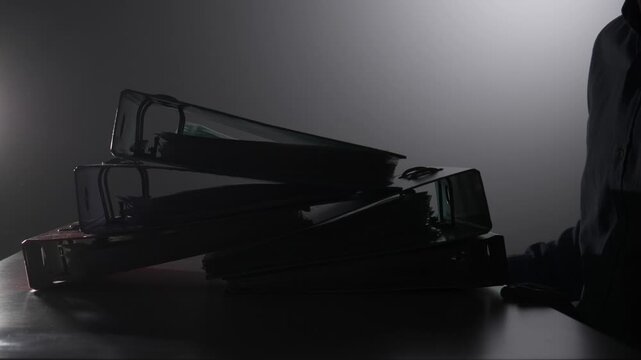 Person's torso and arm entering the frame behind a stack of overflowing binders on a desk. Creating a silhouette against a dark background. Depicting concepts of stress. Burden