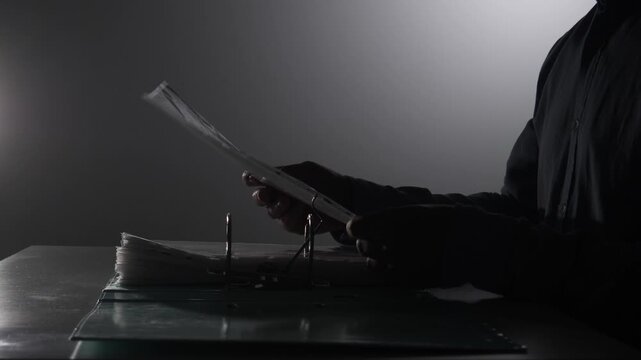 Anonymous person holding and reviewing important evidence files from a dossier, investigating a secret or mystery in a dimly lit, high contrast setting