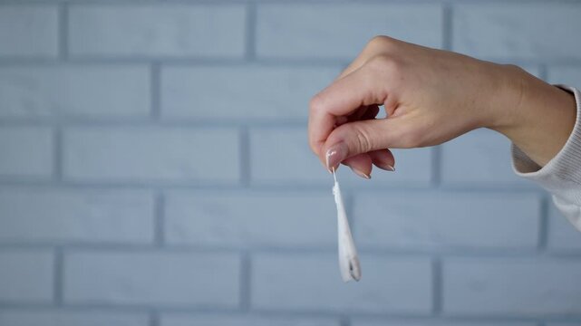 Woman holding a tea bag preparing a hot drink. Female hand dangling a single teabag by its string in front of a blue brick wall background, gently swaying it in preparation for making a hot