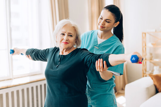 Smiling senior woman doing exercise with help of physical therapist at home in living room