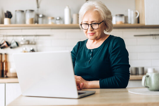 Confident senior woman using laptop for online learning in kitchen copywriter typing on keyboard
