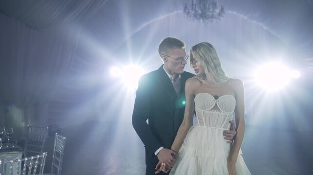 Bride and groom embrace during first dance at wedding reception, illuminated by dramatic blue spotlights and chandelier, romantic atmosphere, elegant white gown and dark suit, soft focus background