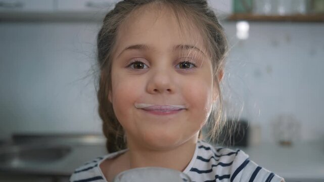 Girl drinking milk from glass in kitchen. Child sips dairy beverage and smiles. Happy kid shows milk mustache on face. Little girl laughs holding milk glass. Healthy child nutrition at home kitchen.