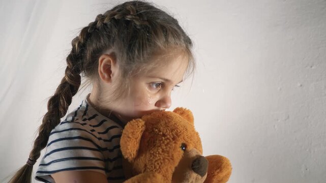 Sad little girl hugs brown teddy bear tightly. Crying child sits alone by white wall. Tear runs down sad face. Lonely kid holds soft toy. Crying girl needs comfort. Bear provides safe feeling.
