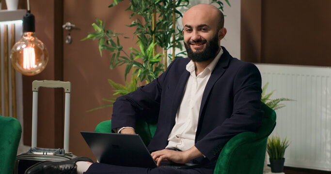 Middle eastern man typing on laptop while relaxing in lobby area and writing speech for global company event. Professional guest travelling on business trip, hotel reception.