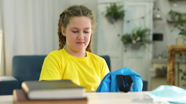 Girl packing school backpack with books at home. Student puts notebook into blue bag. Child prepares backpack for school at desk. Girl organizes books and school supplies. Student getting ready.