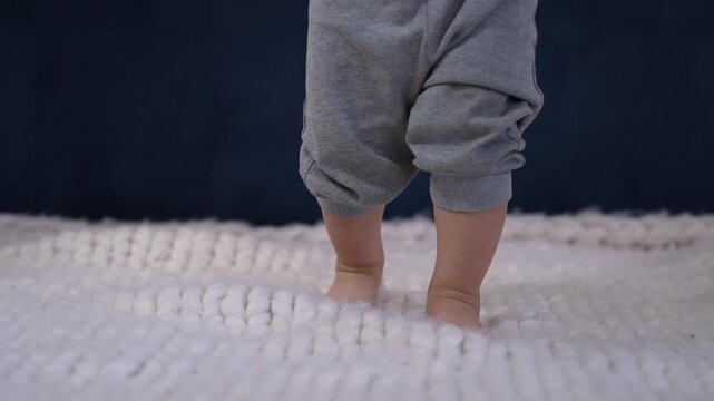 Baby feet standing barefoot on knitted blanket. Toddler bare feet step on soft white wool surface. Small baby leg and tiny toes visible. Infant feet on cozy knit rug. Toddler learns to walk indoors.