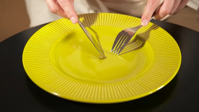 Person cutting a single pea on a plate with cutlery. Conceptual video illustrating diet culture and eating disorders, showing a person cutting a single green pea on a plate with a knife and a fork