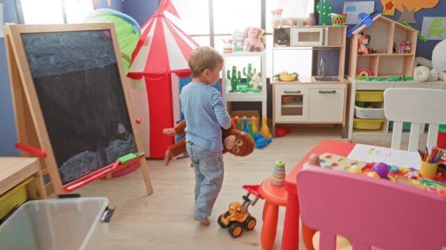 Boy holding a plush monkey with one hand, walking and looking at toys in an indoor playroom inside a building; gentle comfort.