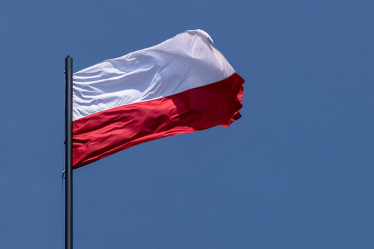 Polska flaga powiewa na wietrze na tle błękitnego nieba. Flaga biało czerwona powiewa na tle czystego, błękitnego nieba. Zawieszona jest na czarnym maszcie, co podkreśla żywy kontrast kolor&oacute;w. 