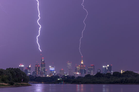 Warsaw, Poland - 02 July 2022: View of powerful lightning bolts striking skyscrapers in the city skyline at night with reflections on the Vistula river.