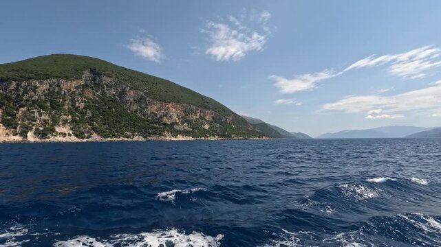 Point of view from a moving boat cruising along the rugged coastline of Ithaca Island in the Ionian Sea, Greece. Deep blue Mediterranean waters, green hills and scenic coastal landscape on a sunny sum
