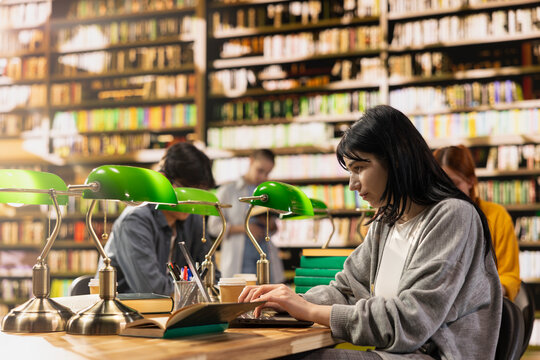 Woman using laptop and open for coursework research at busy study table, surrounded by stacked books and classmates, young adult learning.
